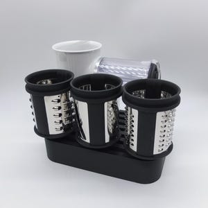 Könnte beinhalten: Ein Küchenhelfer-Set mit drei zylindrischen Reiben mit Edelstahlklingen und schwarzen Rändern, einem weißen Behälter und einem transparenten Kunststoffteil. Das Set steht auf einer schwarzen Basis.