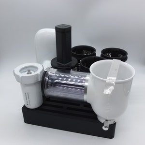 May include: A white electric food grinder with a black handle and multiple attachments. The grinder has a clear plastic cylinder and a white funnel. The base is black with a grid pattern.