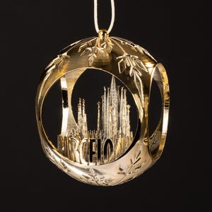 Könnte beinhalten: Goldfarbener Weihnachtsschmuck mit einem Ausschnittdesign, das die Skyline von Barcelona zeigt. Der Schmuck ist kugelförmig mit einer detaillierten architektonischen Darstellung der Stadt. Das Wort "BARCELO" ist sichtbar. Der Schmuck hängt an einer weißen Schnur.