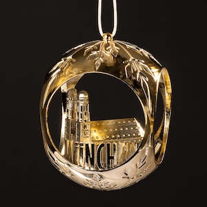 Pode incluir: Um enfeite de Natal dourado com um design detalhado de uma igreja dentro de uma moldura circular. A moldura tem recortes em forma de folha e é suspensa por um fio branco. A palavra "MUNCH" é visível na igreja.