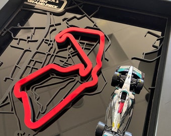 May include: A framed 3D model of the Silverstone Formula 1 racetrack with a miniature silver and green race car on a black background. The frame has a gold plaque that reads "Formula 1 British Grand Prix".
