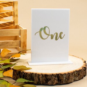 May include: White acrylic table number with a gold 'One' on it. The number is on a wooden stand.