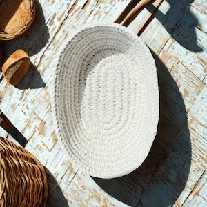 Puede incluir: Vista cenital de una cesta ovalada blanca tejida, vacía, sobre una superficie de madera clara y envejecida. También se ven dos cestas redondas más pequeñas y rebanadas de pan.