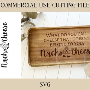 Puede incluir: Una bandeja de servir de madera con el texto "What do you call cheese that doesn't belong to you? Nacho Cheese" grabado en ella. La bandeja está sobre una superficie blanca con una planta verde en el fondo.
