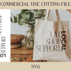 May include: A white tote bag with the text "Eat Shop Support Local" printed in black. The bag is being held by a person's hand.
