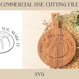 May include: A wooden cutting board with the text "Bake it till you make it." engraved on it. The board is round and has a handle. A small plant is in a pot in the background.