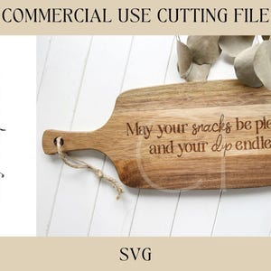 Op de afbeelding: Houten snijplank met handvat, met de gegraveerde tekst: "May your snacks be plentiful and your dip endless." De plank heeft een natuurlijke houtnerf en een jute touwtje om op te hangen. De woorden "COMMERCIAL USE CUTTING FILE" en "SVG" zijn ook zichtbaar.