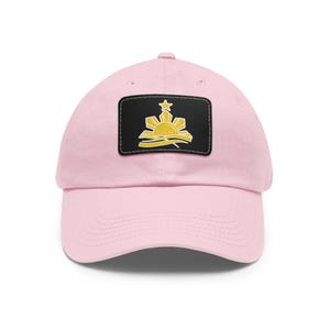 Peut inclure: Casquette de baseball rose clair avec un écusson rectangulaire noir. L'écusson présente un motif de soleil doré avec une étoile et des rayons. La casquette est en tissu doux et a une visière incurvée.