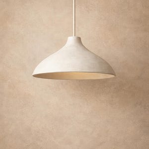 May include: A white, dome-shaped pendant light fixture hanging from a white cord. The light has a smooth, matte finish and a warm, inviting glow emanating from the underside. The background is a textured, neutral-toned wall.