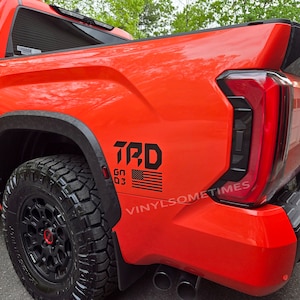 May include: A red pickup truck with a black TRD decal on the side. The decal includes an American flag and the text "GO 03".