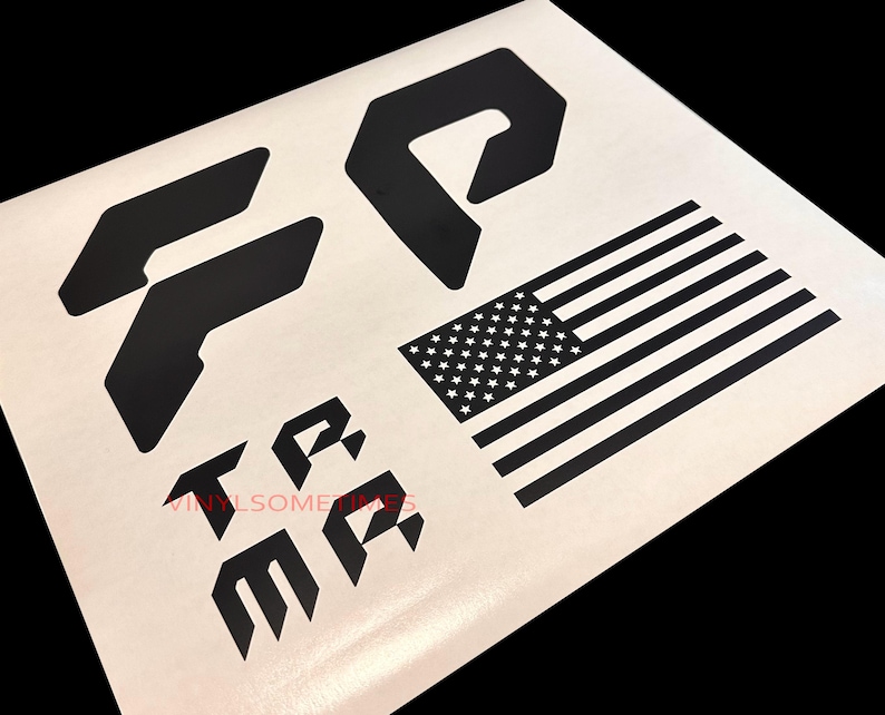 K&ouml;nnte beinhalten: Schwarz-wei&szlig;e Vinyl-Aufkleber mit den Buchstaben "TR" und "MA" in einem stilisierten Design, einer schwarz-wei&szlig;en amerikanischen Flagge und zwei geometrischen Formen.