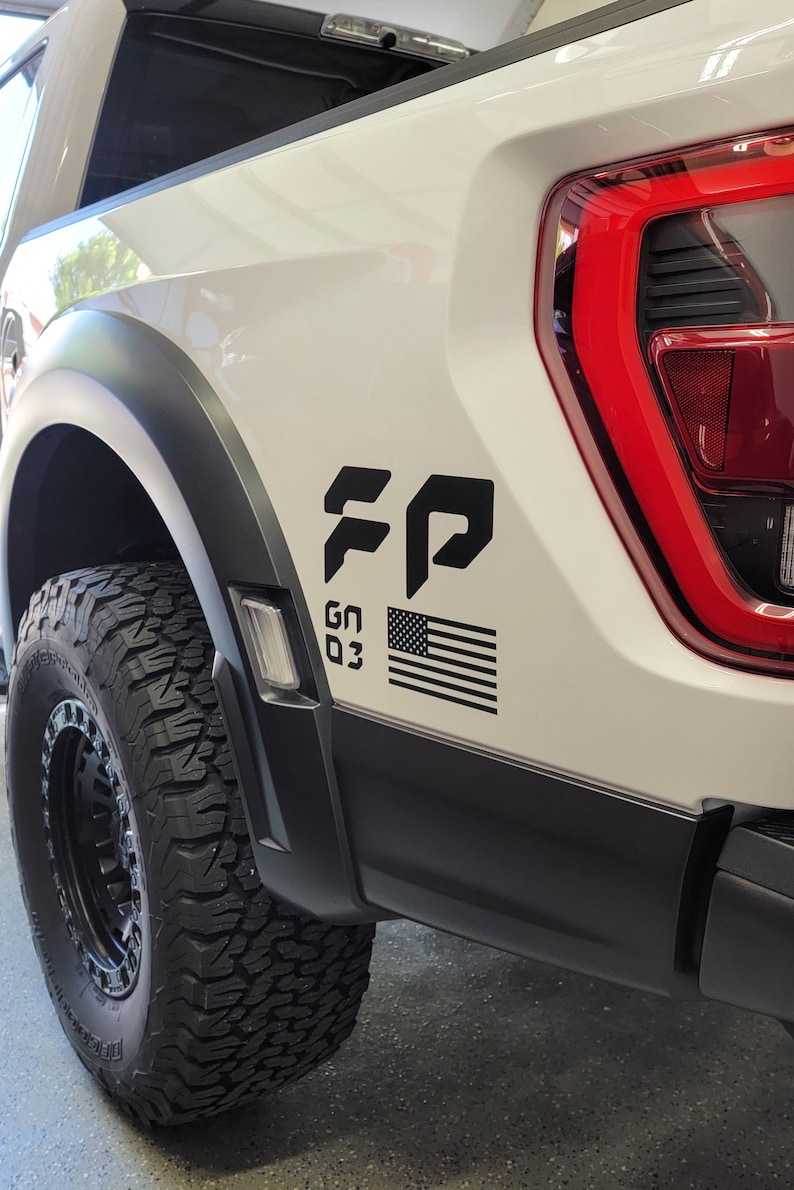 May include: White pickup truck with a black "FP" decal, a small American flag, and the text "GN 03" on the side. The truck has black fender flares and a black rear bumper.