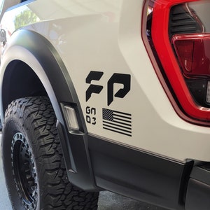 May include: White pickup truck with a black "FP" decal, a black and white American flag decal, and the text "GN 03" on the side. The truck has a black tire and a black wheel.