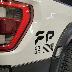 May include: A white pickup truck with a black "FP" logo, an American flag, and "GN 03" text on the side. The truck has a black tire and a red taillight.