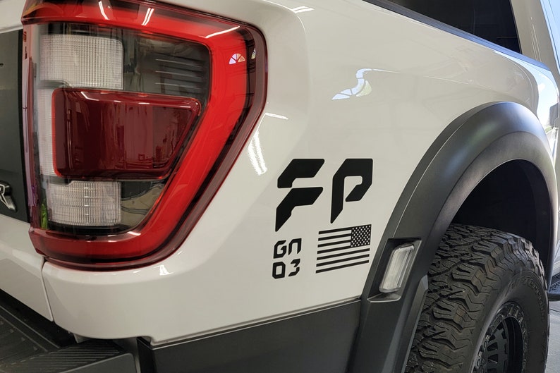 May include: A white pickup truck with a black "FP" logo, an American flag, and "GN 03" text on the side. The truck has a black tire and a red taillight.