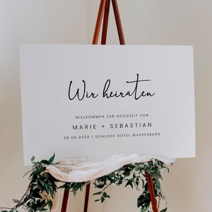 Puede incluir: Un cartel blanco con texto negro en un caballete. El cartel dice "Wir heiraten", que se traduce como "Nos casamos". El cartel también incluye los nombres Marie y Sebastian, la fecha 28-08-2026 y la ubicación Schloss Hotel Wasserburg.