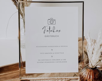 May include: A white sign in a gold frame with the text "Fotobox Gästebuch" and instructions for using a photo booth. The sign is sitting on a wooden surface with a brown vase and dried flowers.