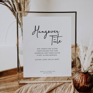 Könnte beinhalten: Ein weißes Schild mit schwarzem Text in einem goldenen Rahmen, auf dem "Hangover T"." steht.  Der Text darunter lautet "Wir haben eine kleine Verpflegung f"."r eure Heimreise und einen guten Start in den Morgen.  Sch"."n, dass ihr da wart.  Marie & Sebastian 28-08-2026".