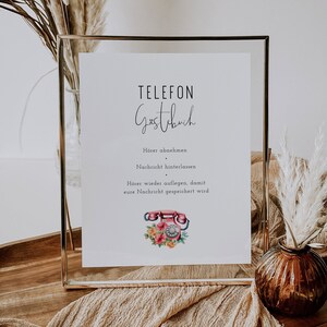 Könnte beinhalten: Ein weißes Schild mit goldenem Rahmen, auf dem in schwarzer Schrift "TELEFON Gastbuch" steht. Das Schild enthält auch Anweisungen zur Verwendung eines antiken Drehwahltelefons als Gästebuch: "Hörer abnehmen, Nachricht hinterlassen. Hörer wieder auflegen, damit eure Nachricht gespeichert wird." Das Schild ist mit einem antiken Drehwahltelefon mit rosa und roten Blumen illustriert.