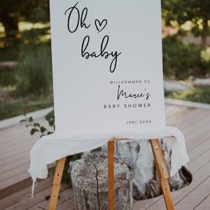 Puede incluir: Un cartel blanco con texto negro que dice "Oh baby" con un s&iacute;mbolo de coraz&oacute;n. El cartel tambi&eacute;n dice "Willkommen zu Marie's Baby Shower Juni 2026". El cartel est&aacute; apoyado en un caballete de madera.