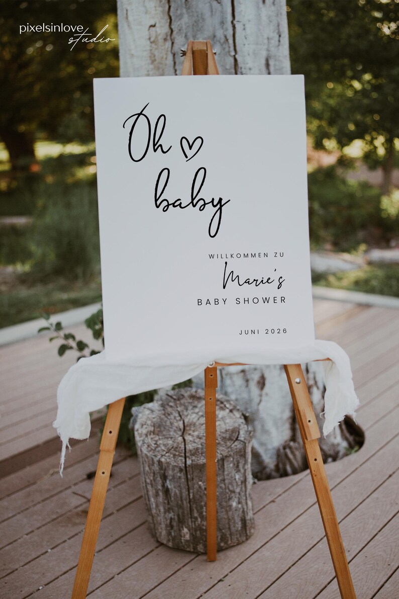 Puede incluir: Un cartel blanco con texto negro que dice "Oh baby" con un s&iacute;mbolo de coraz&oacute;n. El cartel tambi&eacute;n dice "Willkommen zu Marie's Baby Shower Juni 2026". El cartel est&aacute; apoyado en un caballete de madera.