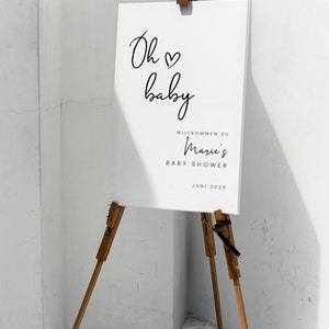 Puede incluir: Un cartel blanco con caligraf&iacute;a negra que dice "Oh baby" con un s&iacute;mbolo de coraz&oacute;n. El cartel tambi&eacute;n dice "Willkommen zu Marie's Baby Shower Juni 2026". El cartel est&aacute; en un caballete de madera.
