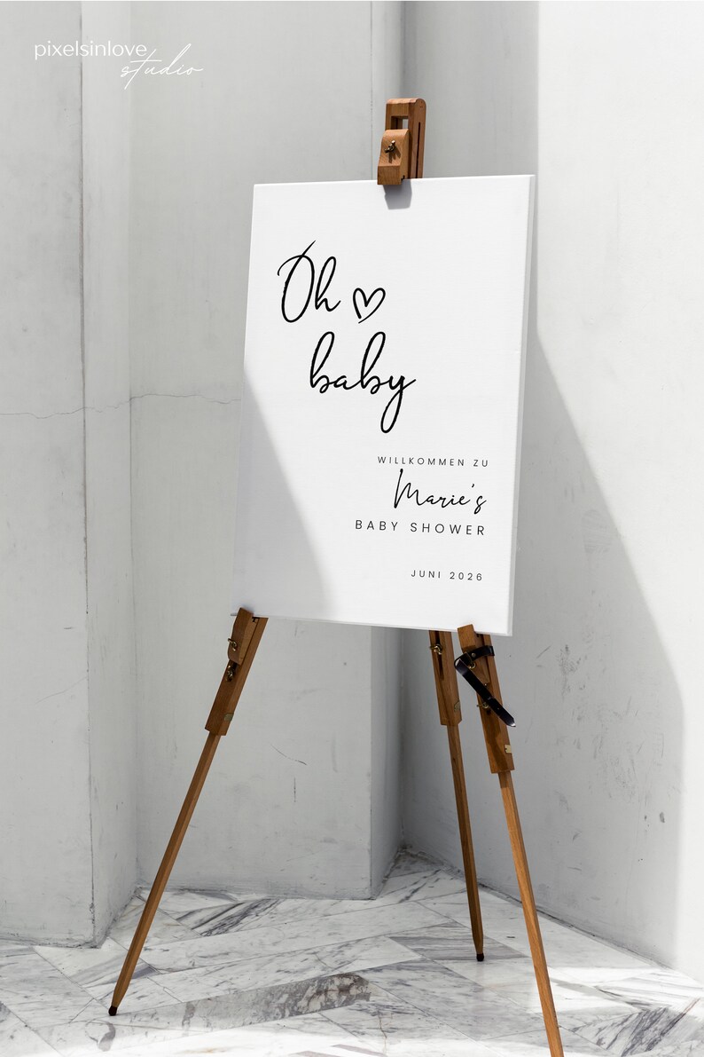 Puede incluir: Un cartel blanco con caligraf&iacute;a negra que dice "Oh baby" con un s&iacute;mbolo de coraz&oacute;n. El cartel tambi&eacute;n dice "Willkommen zu Marie's Baby Shower Juni 2026". El cartel est&aacute; en un caballete de madera.