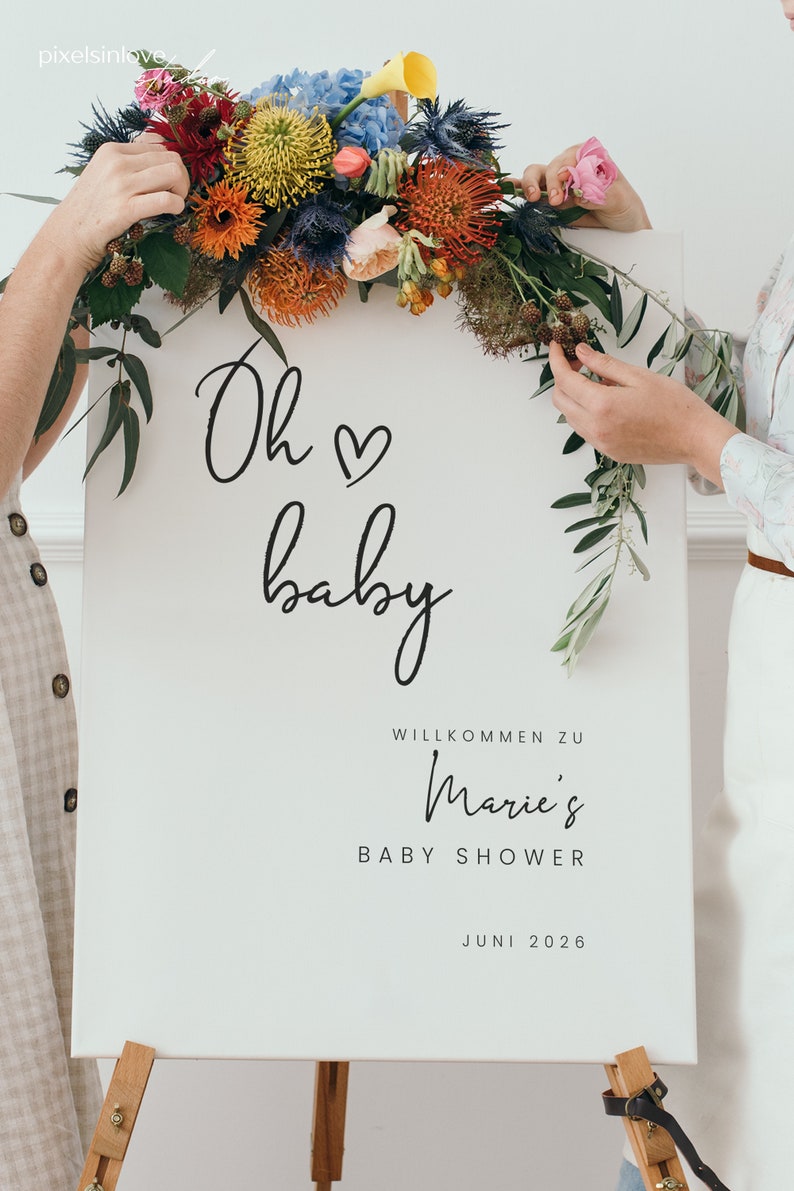 Puede incluir: Un cartel blanco con caligraf&iacute;a negra que dice "Oh baby" con un s&iacute;mbolo de coraz&oacute;n. El cartel tambi&eacute;n dice "Willkommen zu Marie's Baby Shower Juni 2026". El cartel est&aacute; decorado con una guirnalda floral de flores rojas, naranjas, amarillas y azules.