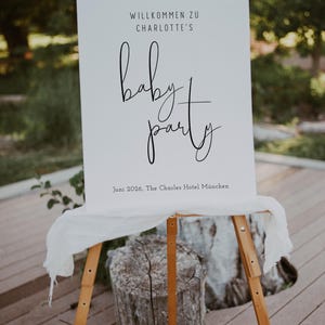Puede incluir: Un cartel blanco con texto negro anunciando una fiesta de beb&eacute; para Charlotte. El cartel dice "Willkommen zu Charlotte's baby party" y "Juni 2026, The Charles Hotel M&uuml;nchen".
