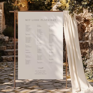 May include: A rectangular white seating chart sign with the text MIT LIEBE PLATZIERT and a list of names, suspended on a copper-colored metal frame stand. A long, draped cream-colored fabric hangs from the right side of the frame.