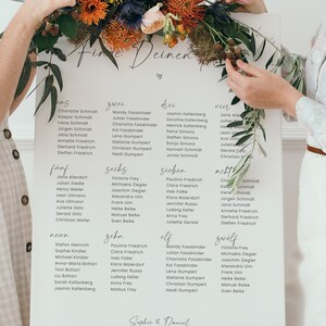 May include: A white card with the words "Finde Deinen" written in black calligraphy. The card is divided into twelve sections, each with a number and a list of names. The card is decorated with a floral arrangement of colourful flowers.