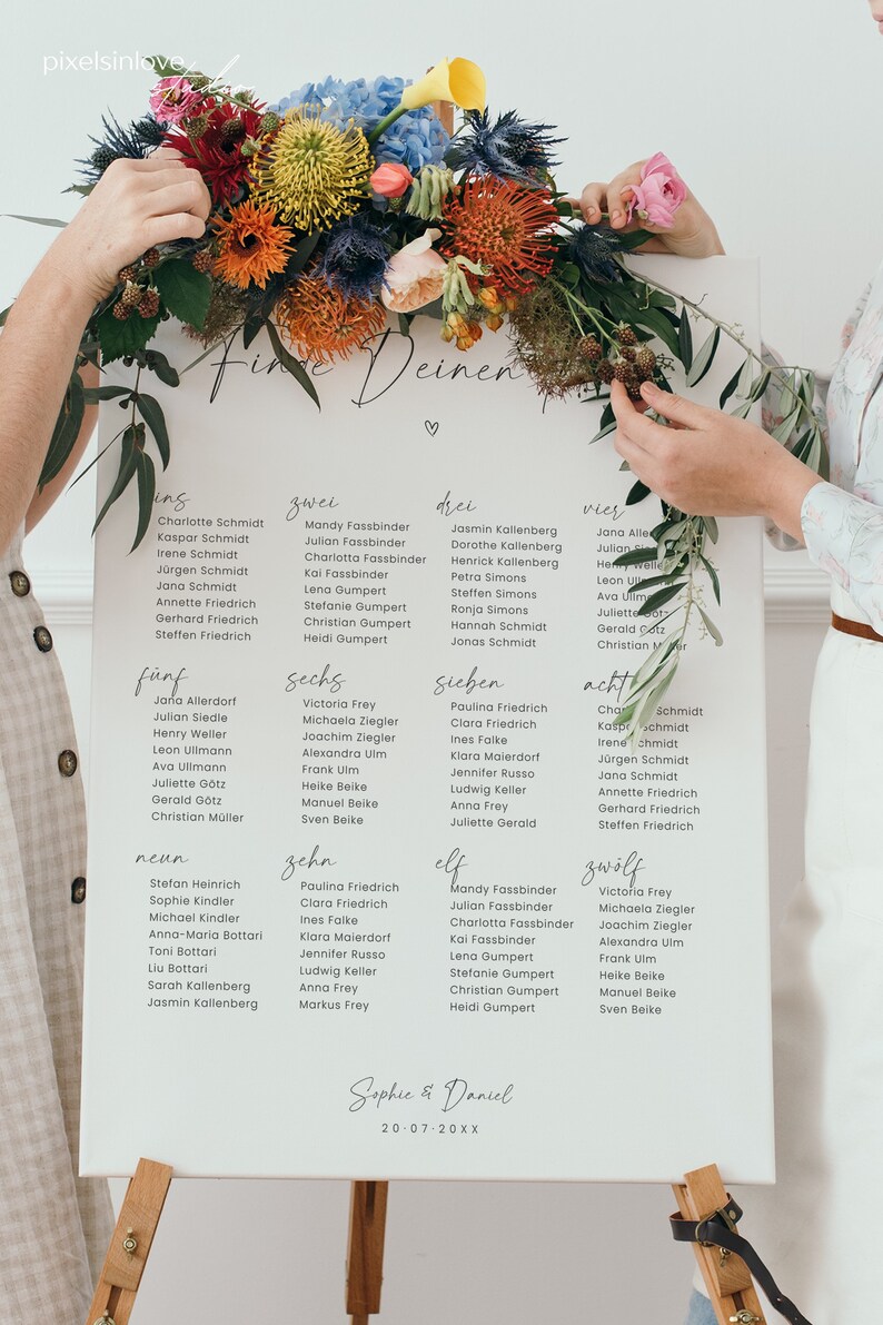 May include: A white card with the words "Finde Deinen" written in black calligraphy. The card is divided into twelve sections, each with a number and a list of names. The card is decorated with a floral arrangement of colourful flowers.