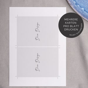 May include: Two white cards with the text "Dein Design" written in black cursive on each card. The cards are on a gray surface with a blue plate in the background. The text "MEHRERE KARTEN PRO BLATT DRUCKEN SELBST ZUSCHNEIDEN" is written in black on a white circle.