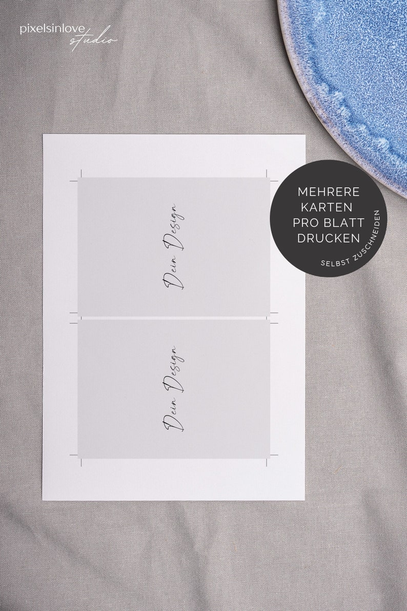 May include: Two white cards with the text "Dein Design" written in black cursive on each card. The cards are on a gray surface with a blue plate in the background. The text "MEHRERE KARTEN PRO BLATT DRUCKEN SELBST ZUSCHNEIDEN" is written in black on a white circle.