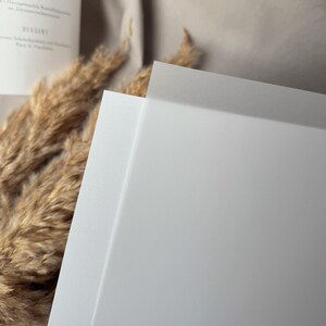 May include: Close-up of white paper, possibly cardstock, with a textured edge. The paper is stacked, with the top sheet slightly angled, revealing a clean, bright surface. Dried ornamental grass and a menu are in the background.
