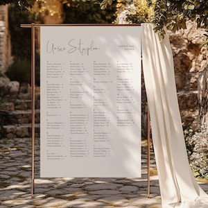 Puede incluir: Un plano de asientos de boda exhibido en un marco de metal color cobre. El gráfico presenta un fondo color crema con caligrafía elegante. El texto "Unser Sitzplan" está en la parte superior, con los nombres de los invitados y los números de mesa debajo.