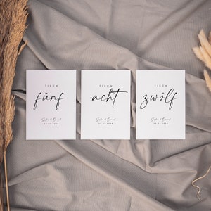 May include: Three white cards with black text on a gray fabric background. The cards are labeled "TISCH f&uuml;nf", "TISCH acht", and "TISCH zw&ouml;lf". Each card also includes the text "Sophie & Daniel" and a date.