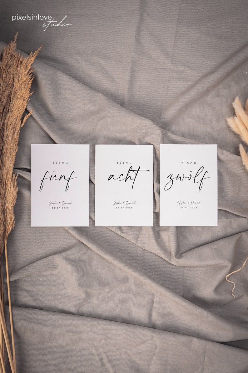 May include: Three white cards with black text on a gray fabric background. The cards are labeled "TISCH f&uuml;nf", "TISCH acht", and "TISCH zw&ouml;lf". Each card also includes the text "Sophie & Daniel" and a date.