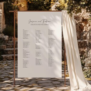 May include: A vertical white wedding seating chart sign with black text reading Jasmin und Fabian and Schloss Wangen Bellermont, displayed on a copper metal frame stand with a draped cream fabric accent hanging from the right side.