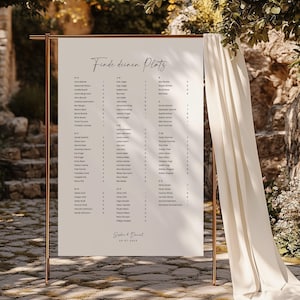 May include: A rectangular white wedding seating chart sign with black text titled Finde deinen Platz, featuring guest names organized alphabetically by letter groups and table numbers, suspended on a copper metal frame with draped fabric.