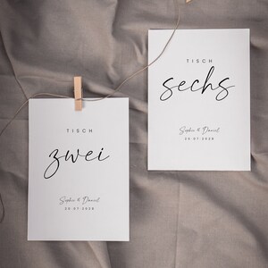 May include: Two white cards with black text on a brown fabric background. The cards are for a wedding and have the table numbers "zwei" and "sechs" written on them. The cards also include the names "Sophie & Daniel" and the date "20.07.2028".