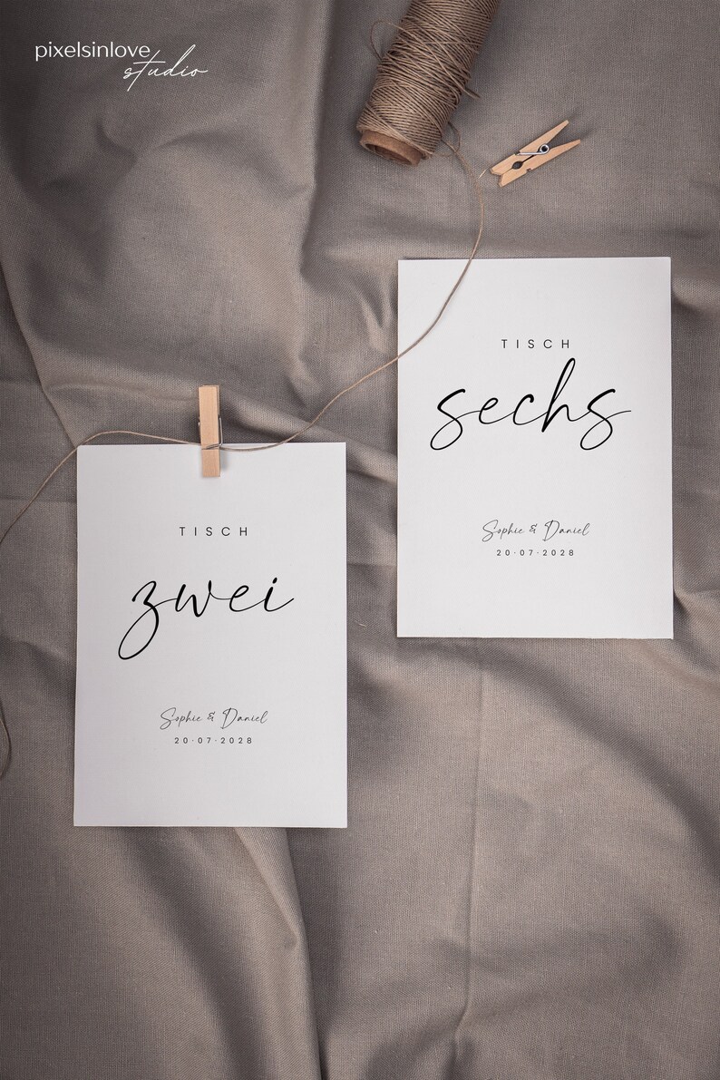 May include: Two white cards with black text on a brown fabric background. The cards are for a wedding and have the table numbers "zwei" and "sechs" written on them. The cards also include the names "Sophie & Daniel" and the date "20.07.2028".