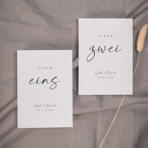 May include: Two white cards with black text on a gray fabric background. The cards are table number signs for a wedding. The first card says "Tisch eins" and the second card says "Tisch zwei". Both cards include the names "Sophie & Daniel" and the date "20.07.2028".