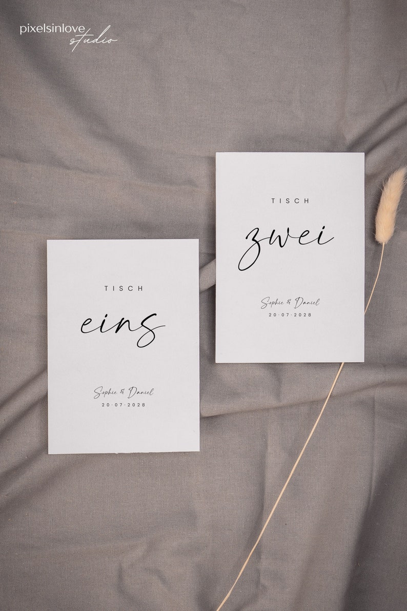 May include: Two white cards with black text on a gray fabric background. The cards are table number signs for a wedding. The first card says "Tisch eins" and the second card says "Tisch zwei". Both cards include the names "Sophie & Daniel" and the date "20.07.2028".