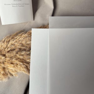 May include: A close-up shot of a menu card with German text, including the word "DESSERT". The card is white with a simple, elegant design. The background features a neutral-toned fabric and dried ornamental grass.