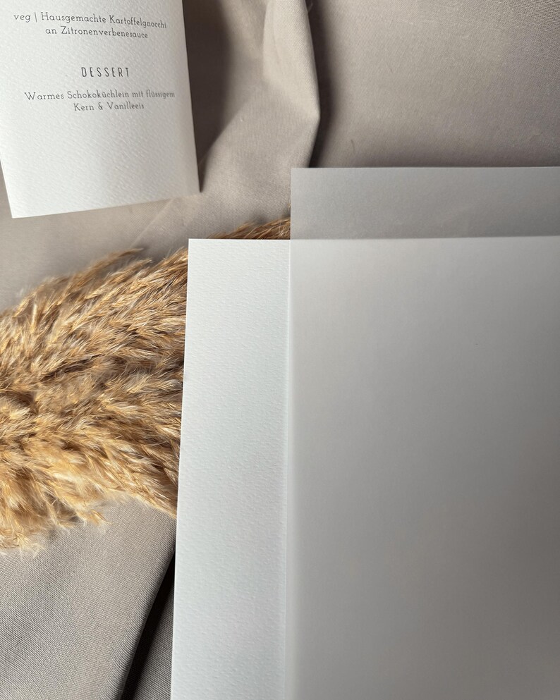 May include: A close-up shot of a menu card with German text, including the word "DESSERT". The card is white with a simple, elegant design. The background features a neutral-toned fabric and dried ornamental grass.