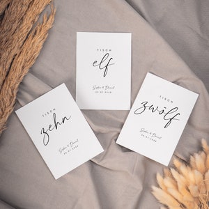 May include: Three white table cards with black calligraphy. The cards are labeled "TISCH elf", "TISCH zehn", and "TISCH zw&ouml;lf". Each card includes the text "Sophie & Daniel" and a date.