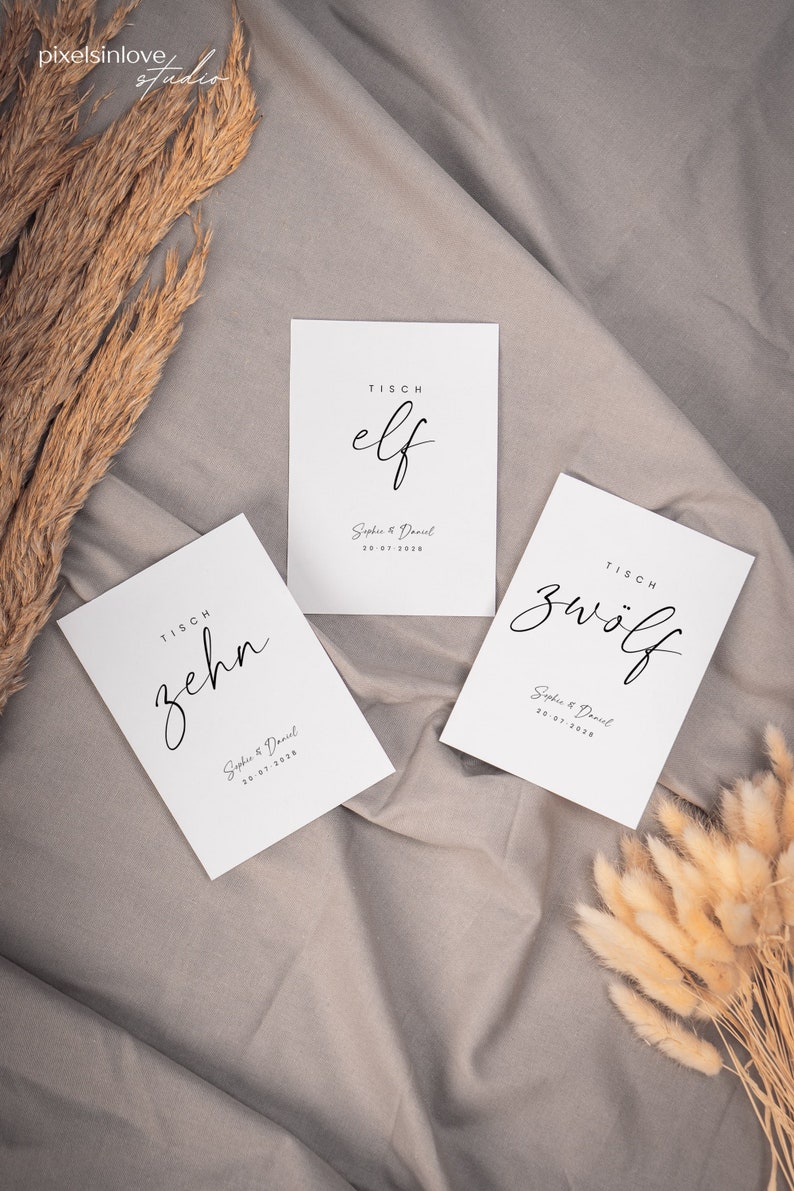 May include: Three white table cards with black calligraphy. The cards are labeled "TISCH elf", "TISCH zehn", and "TISCH zw&ouml;lf". Each card includes the text "Sophie & Daniel" and a date.