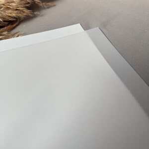 May include: A close-up shot of two white sheets of paper, one slightly overlapping the other. The paper has a smooth, matte finish. The background is a light gray fabric, with a hint of dried ornamental grass in the upper left corner.