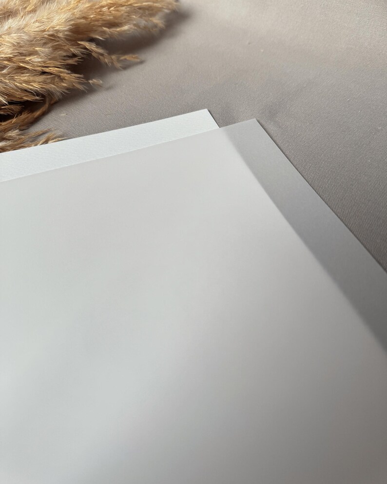 May include: A close-up shot of two white sheets of paper, one slightly overlapping the other. The paper has a smooth, matte finish. The background is a light gray fabric, with a hint of dried ornamental grass in the upper left corner.
